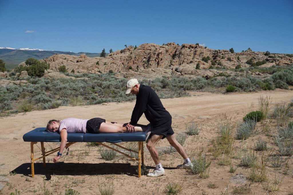 Physical therapist providing leg treatment to athlete on massage table in desert terrain with rocky hills