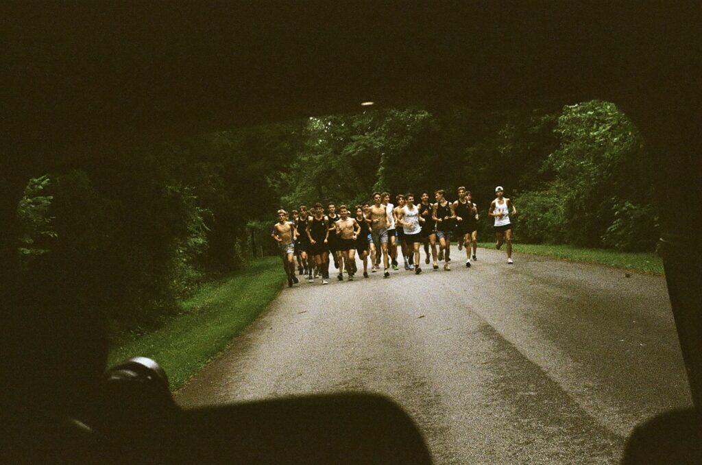 Group of high school runners mid-run on a wooded road, viewed from a following vehicle.