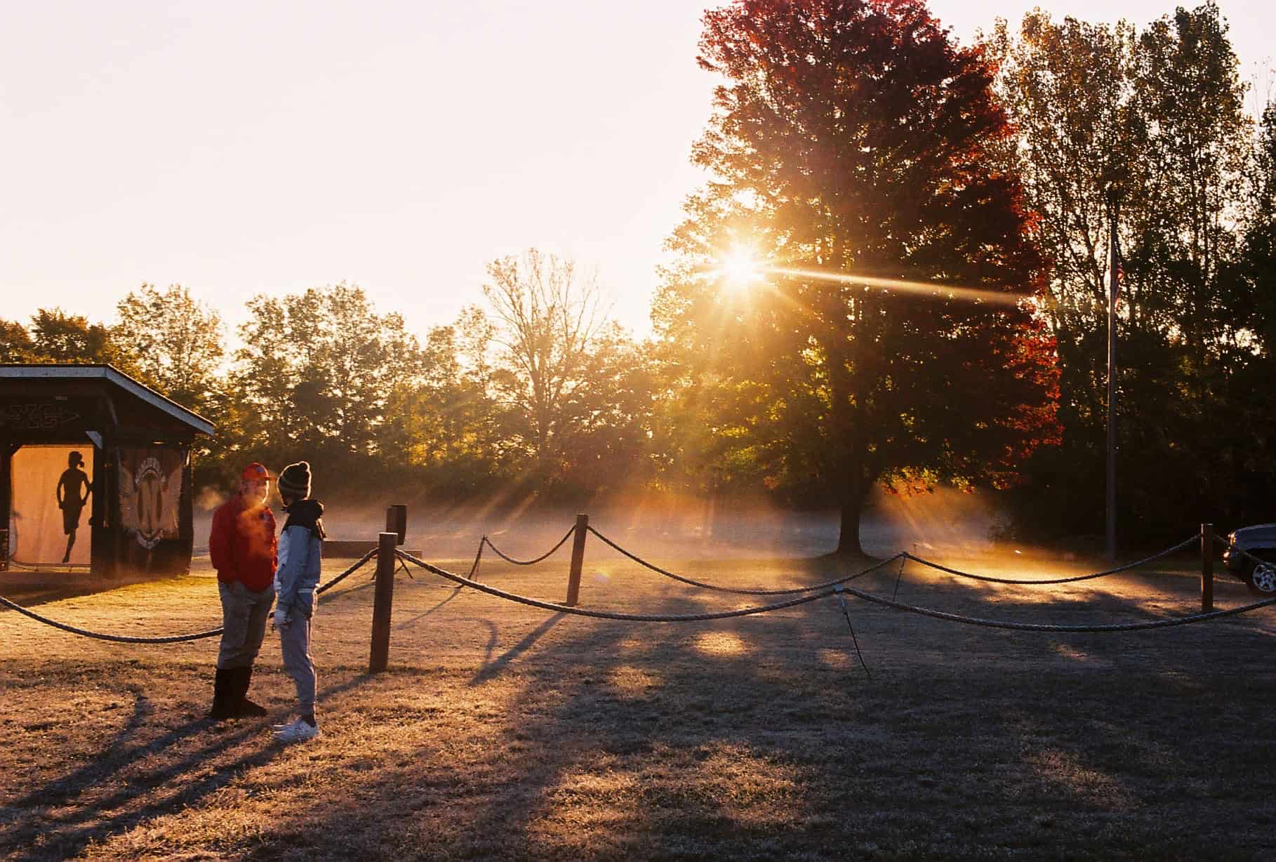 Two people standing near a frosty cross country course at sunrise with sun rays through a red tree