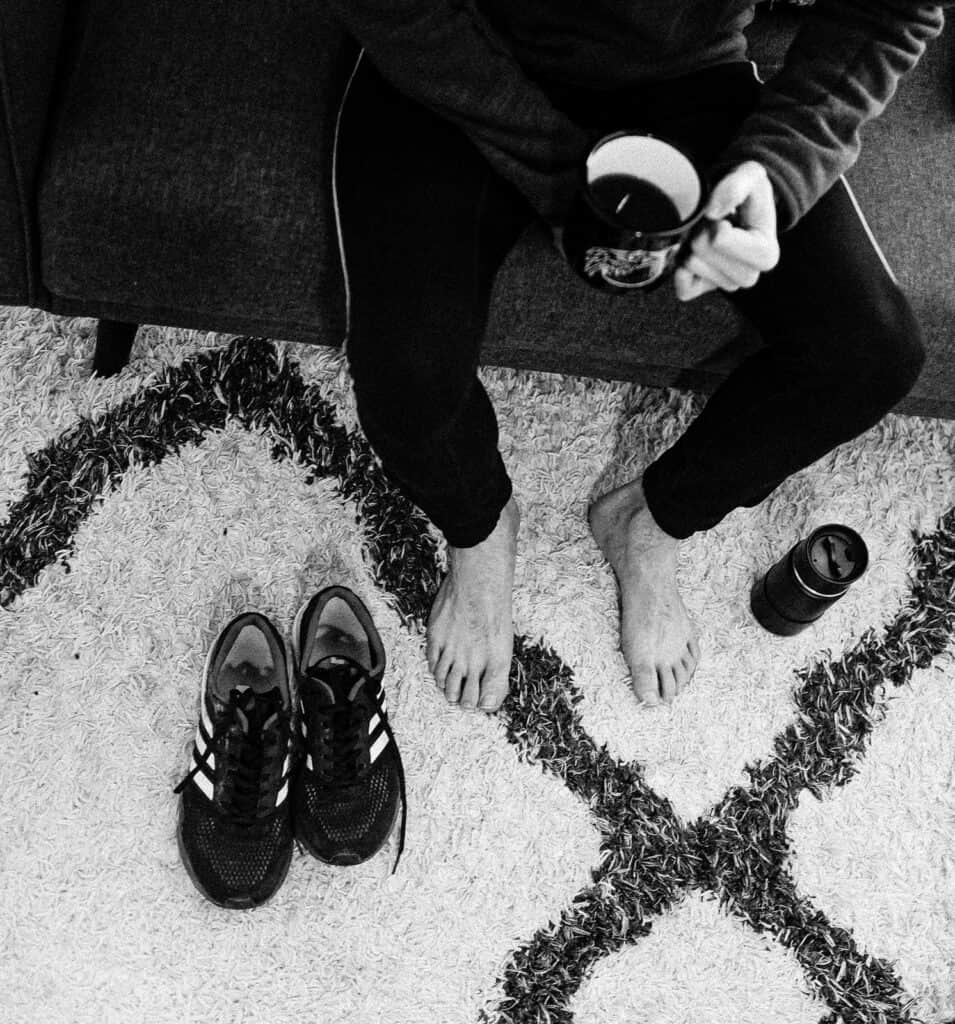 Person sitting barefoot on a rug with running shoes and coffee, seen from above.