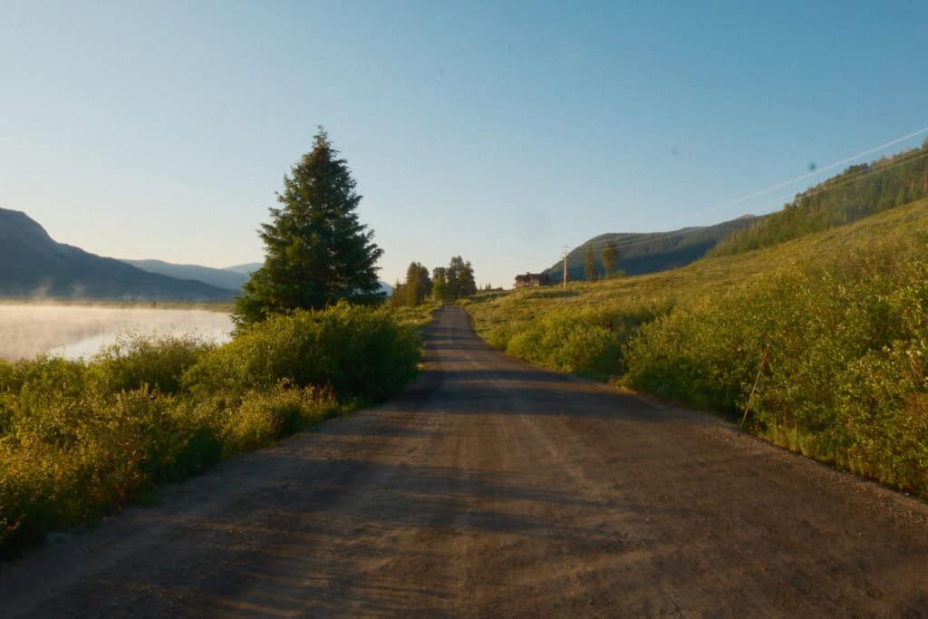 A dirt road leads into the distance between early morning trees and misty water with mountains in the background