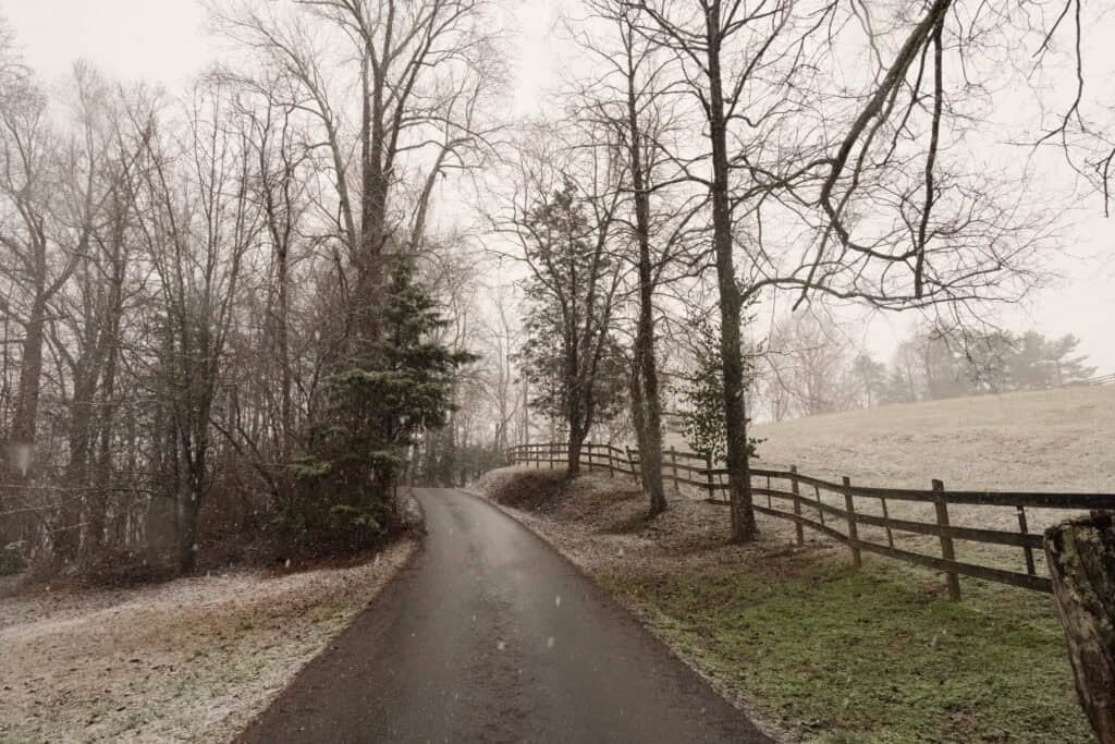 Narrow country road winding uphill through a snowy rural landscape with bare trees and wooden fences