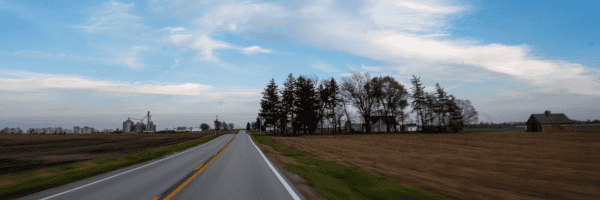 A two-lane country road stretching toward the horizon with farmland and scattered trees under a partly cloudy evening sky